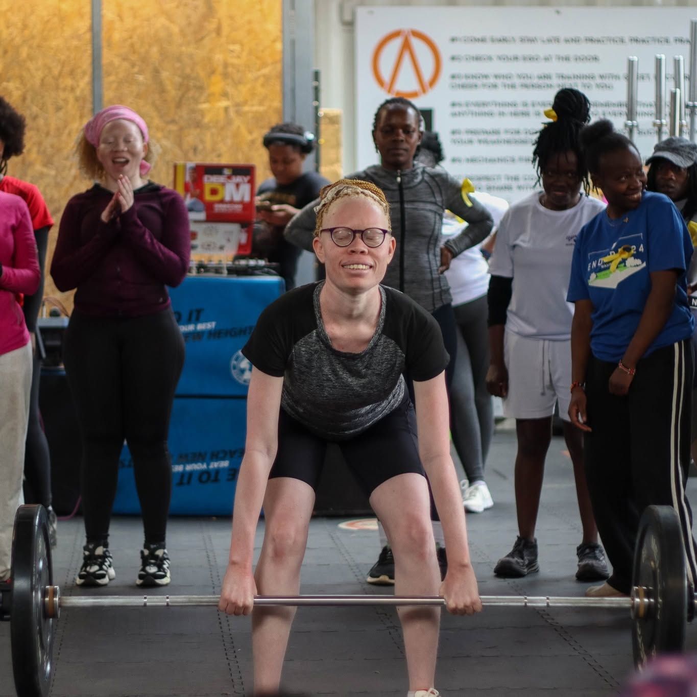 A woman with albinism lifts a loaded barbell in a gym while other athletes cheer behind her.